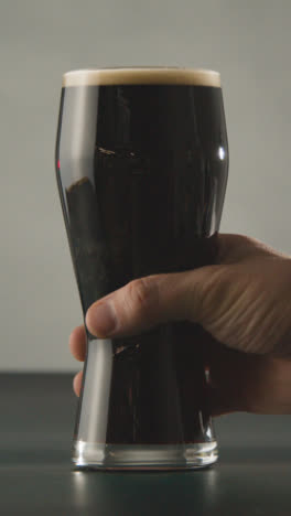 Vertical-Video-Of-Person-Picking-Up-Pint-Of-Irish-Stout-In-Glass-Against-Grey-Studio-Background-To-Celebrate-St-Patricks-Day